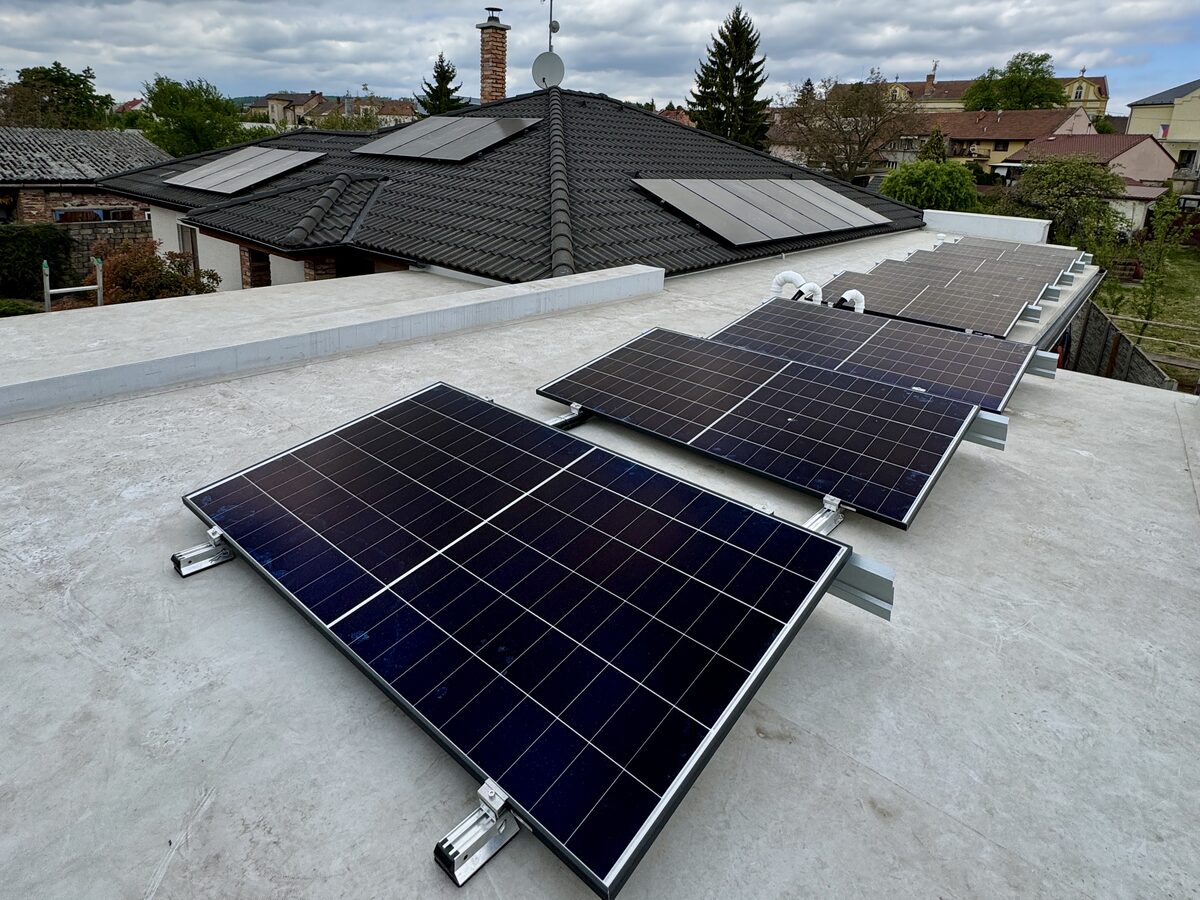 Fotovoltaické panely na ploché střeše rodinného domu — hliníková nosná konstrukce, další panely na sedlové střeše v pozadí